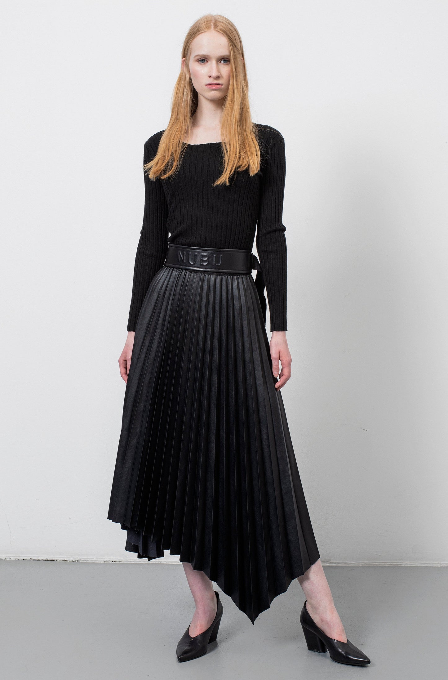 Ginger female model wearing a knit black top with long sleeves, a black leather embossed belt and a vegan leather asymmetrical pleated skirt with heeled black leather sandals.